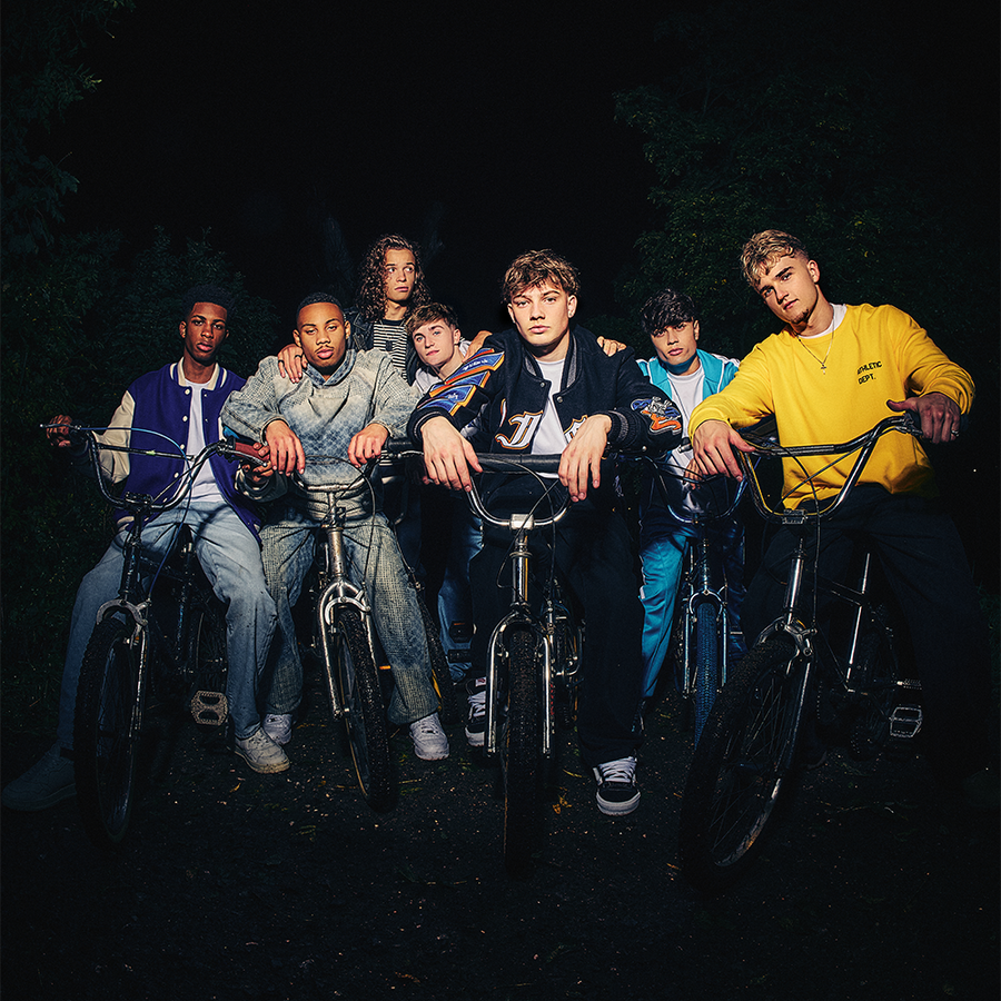 A group of seven young men posing with bicycles in a dark outdoor setting at night, illuminated by dramatic low-key lighting. They are dressed in casual streetwear, including a yellow sweatshirt, blue jacket, and various denim and leather outfits.