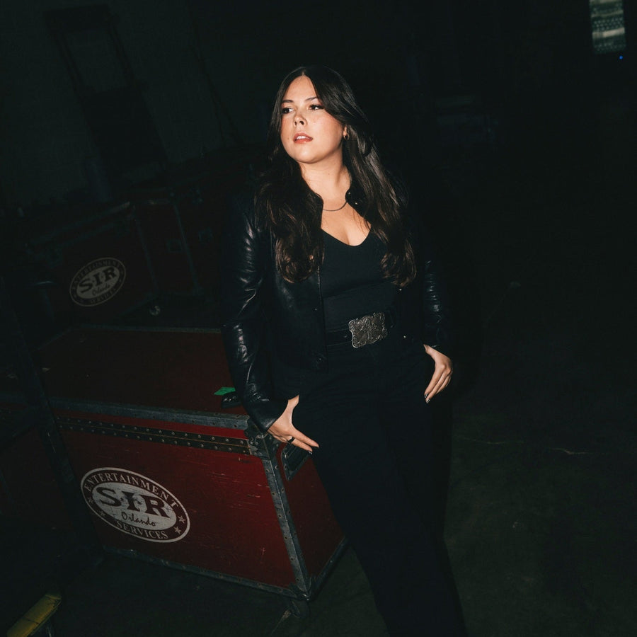 ALT POP STAR GIGI PEREZ standing next to a red and black suitcase with 'STIR' branding in a dark setting